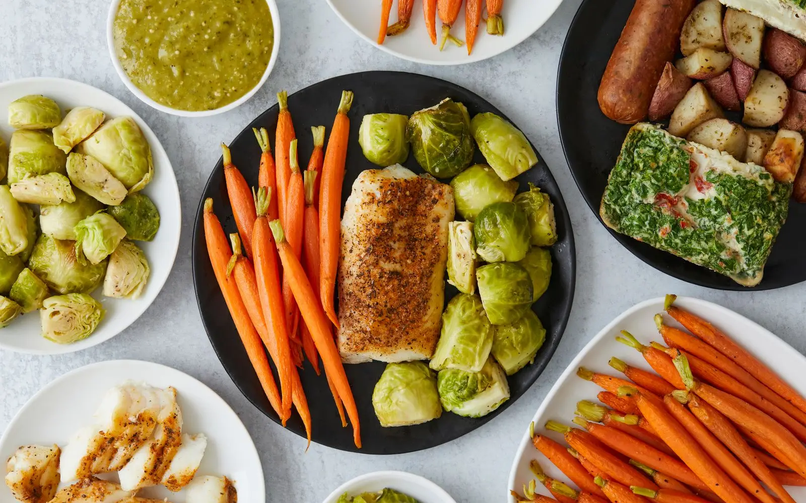 Top view of a dark plate with a fillet of white fish, carrots and Brussels sprouts, with more vegetable dishes around it.