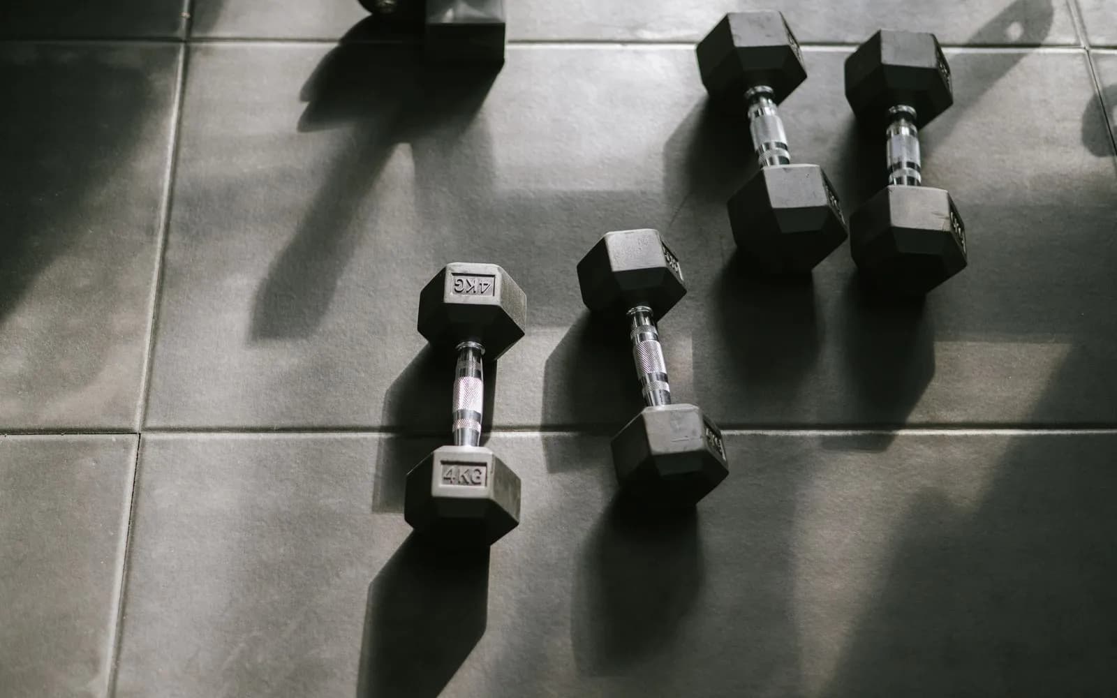 Four small black hexagonal dumbbells lying on a grey gym floor in sunlight.