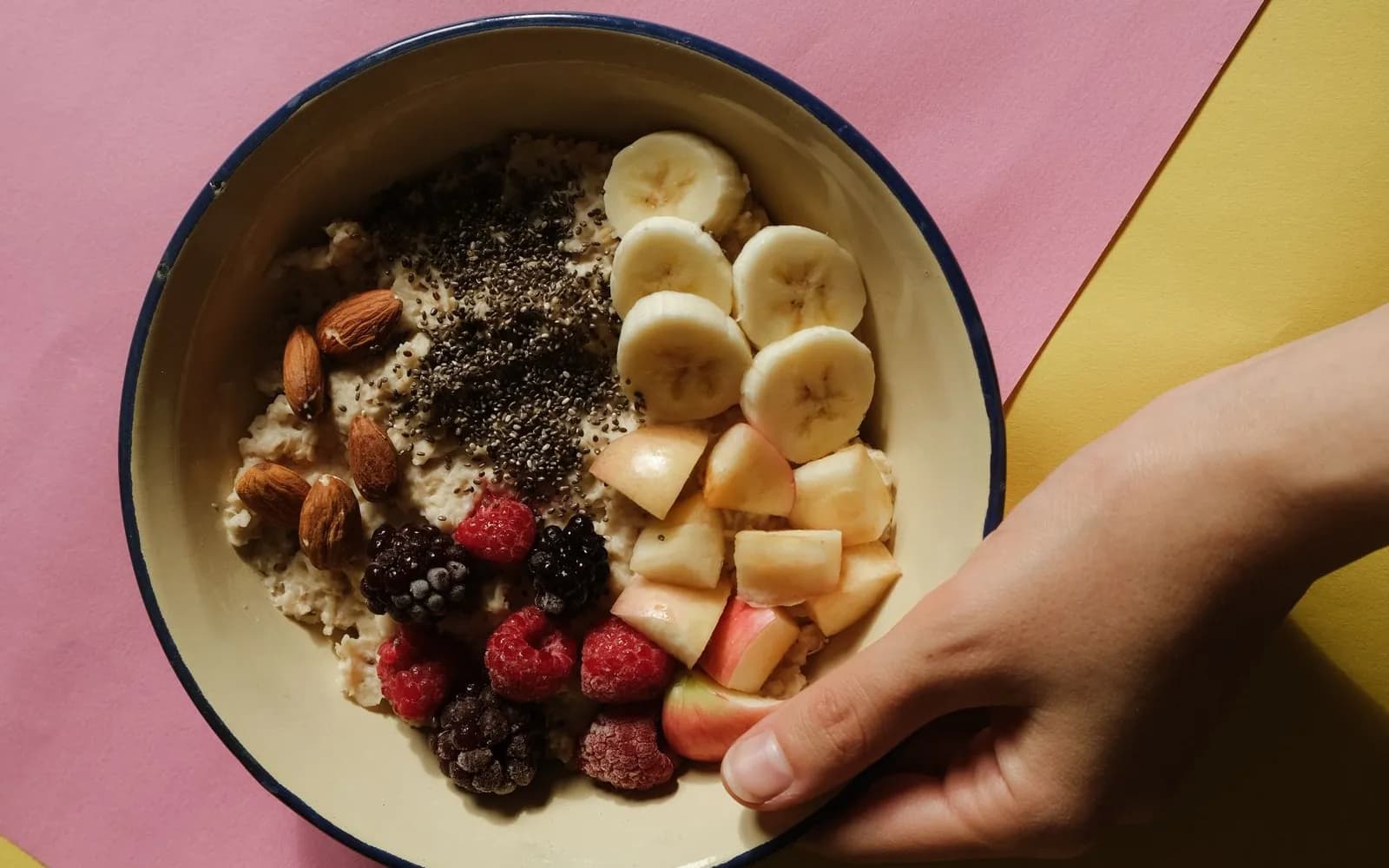 A hand holding a bowl of porridge topped with sliced banana, chopped apple, berries, almonds and chia seeds, on pink and yellow paper
