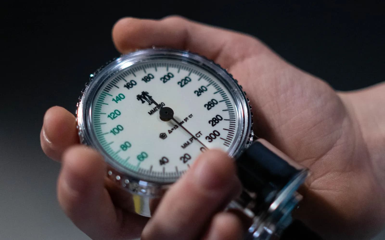 A hand holding the round dial of a blood pressure gauge, with its scale marked from 0 to 300, against a dark background