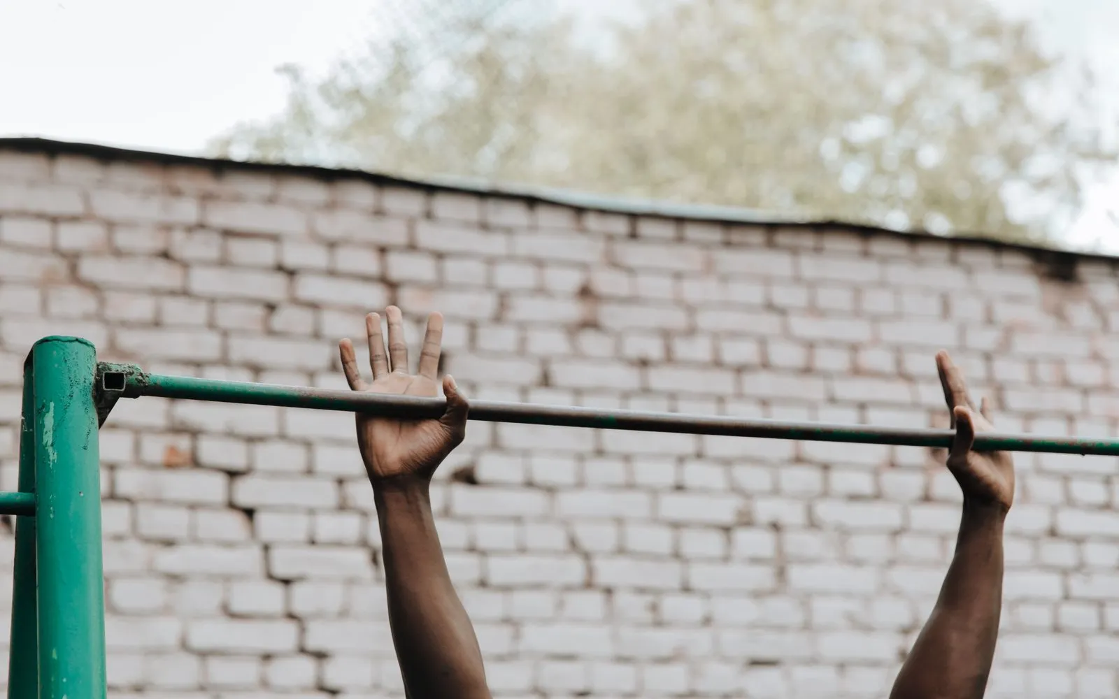 Two hands holding an outdoor metal pull-up bar in front of a pale brick wall
