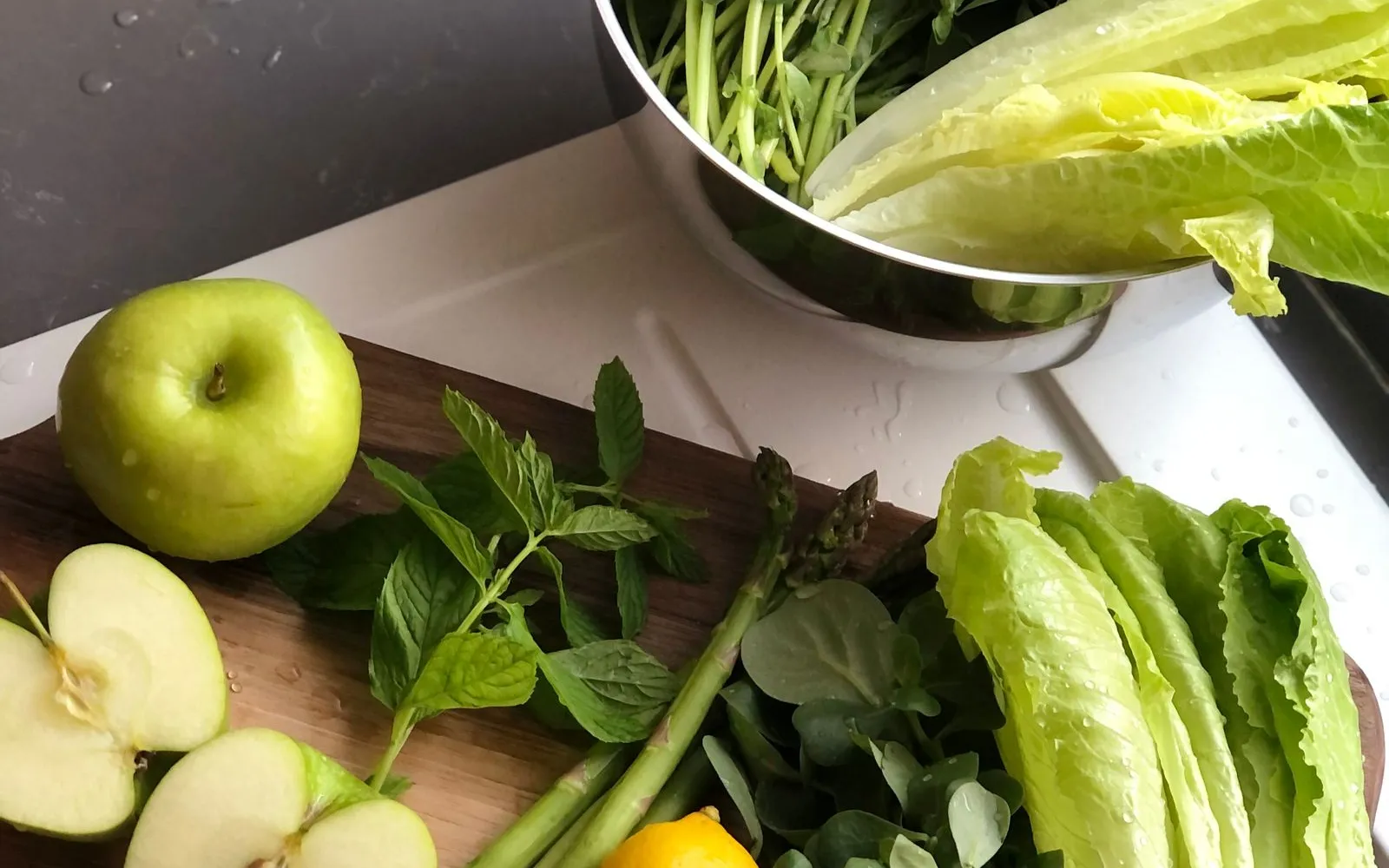 Green apples, lemons, asparagus, mint and romaine lettuce on a wooden board, with a steel bowl of fresh greens behind.