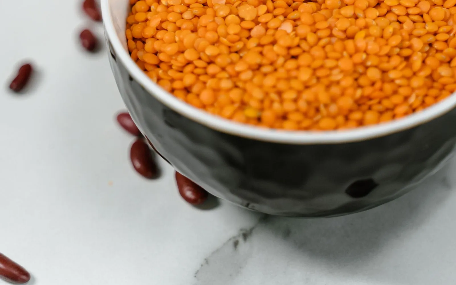 A bowl of dry red lentils on a white marble worktop, with red kidney beans scattered around it.