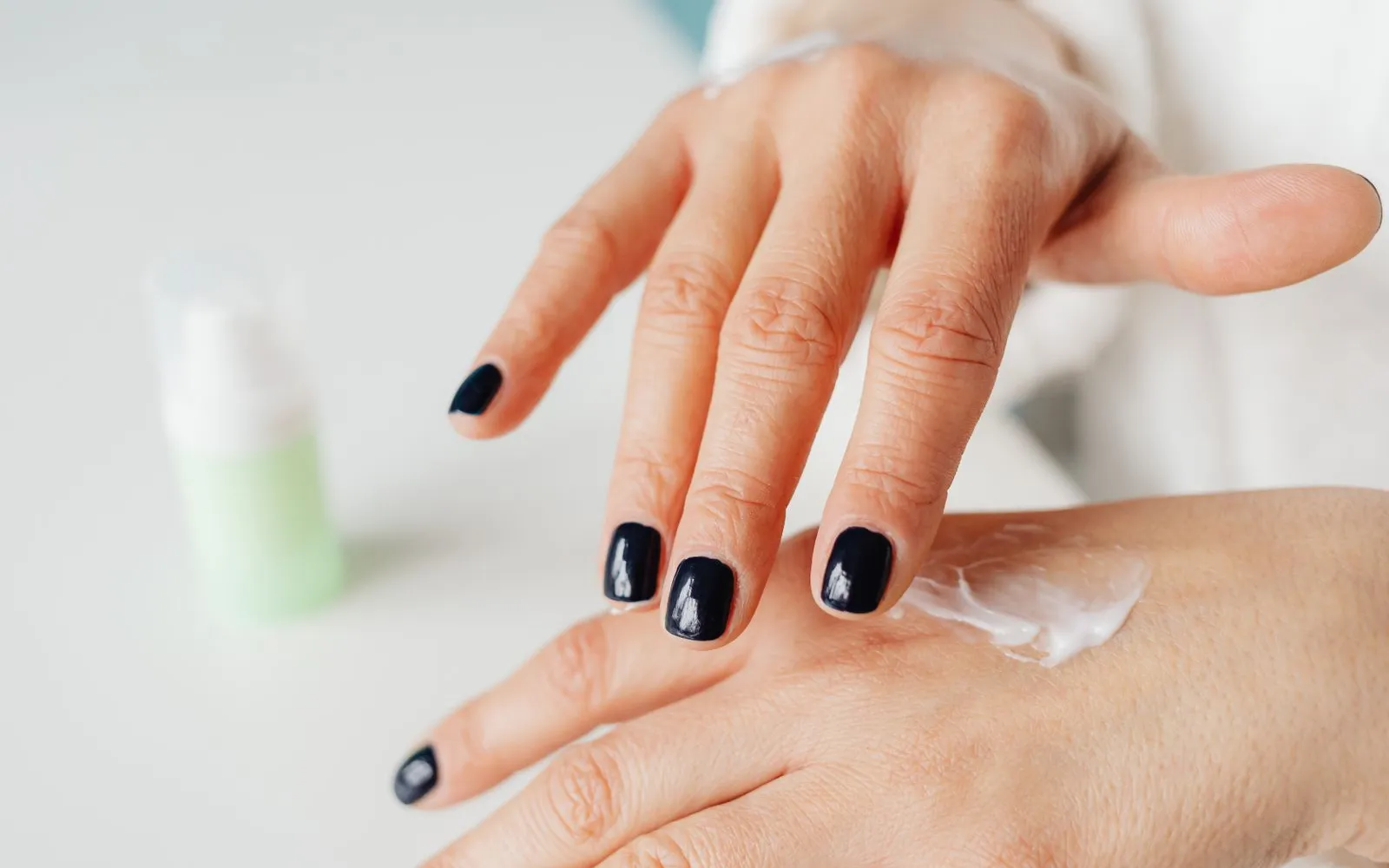 Close-up of hands rubbing white cream into the back of one hand, with a blurred unlabelled bottle behind