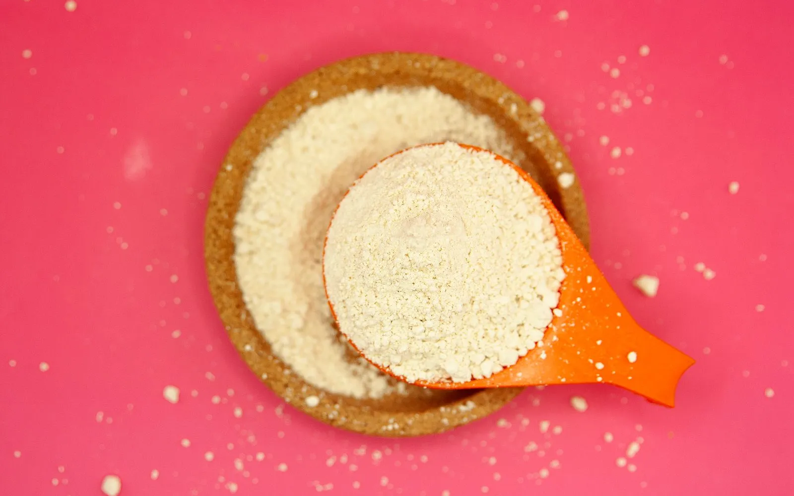 A scoop of pale powder resting in a cork bowl of the same powder on a pink background