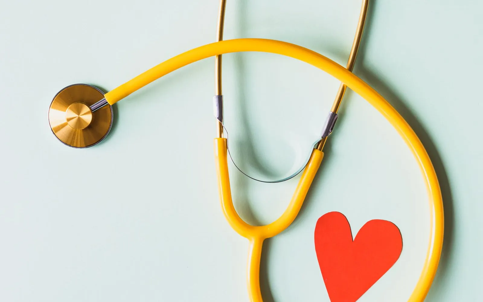 A yellow stethoscope curled around a red paper heart on a pale background.