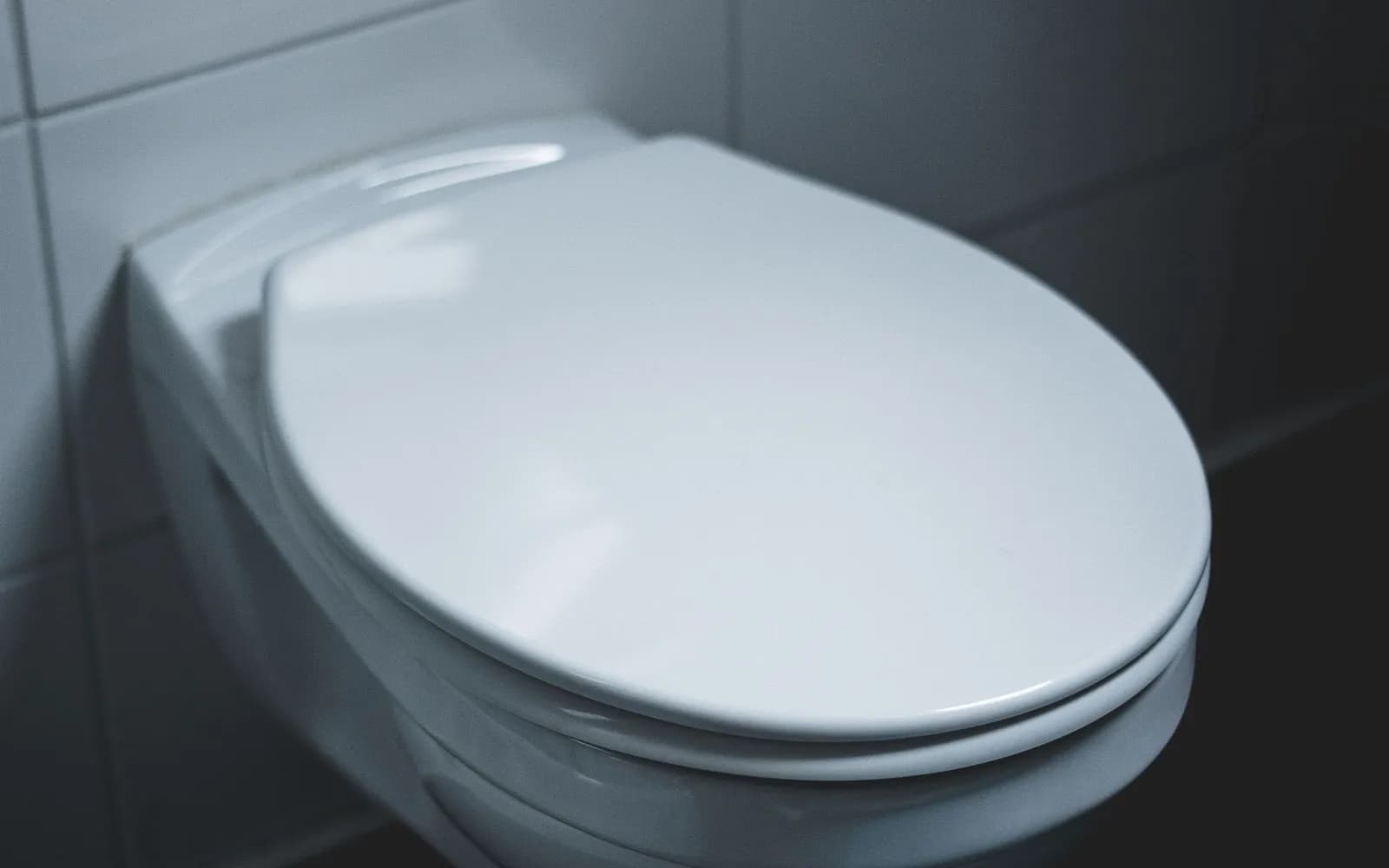 A white wall-hung toilet with the lid down in a dimly lit tiled bathroom