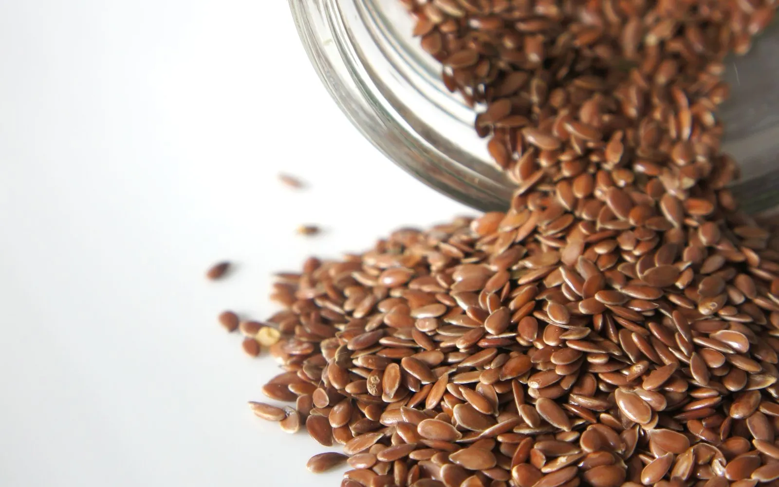 Brown flax seeds spilling from a tipped glass jar onto a white surface