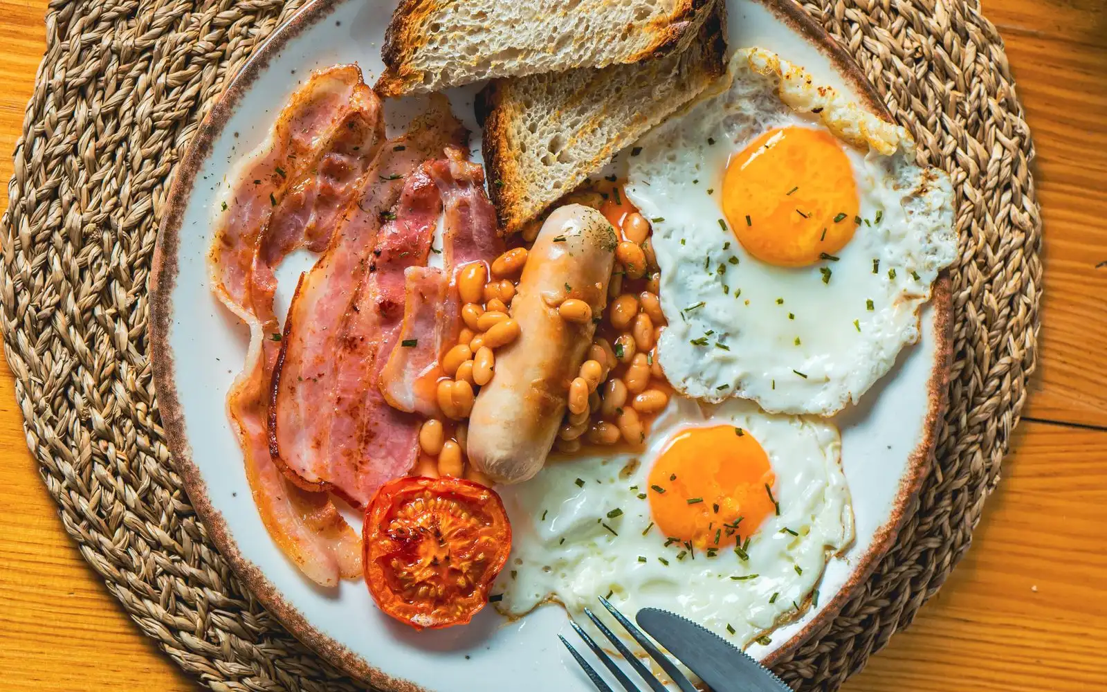 A cooked breakfast of bacon, a sausage, baked beans, two fried eggs, grilled tomato and toast on a white plate