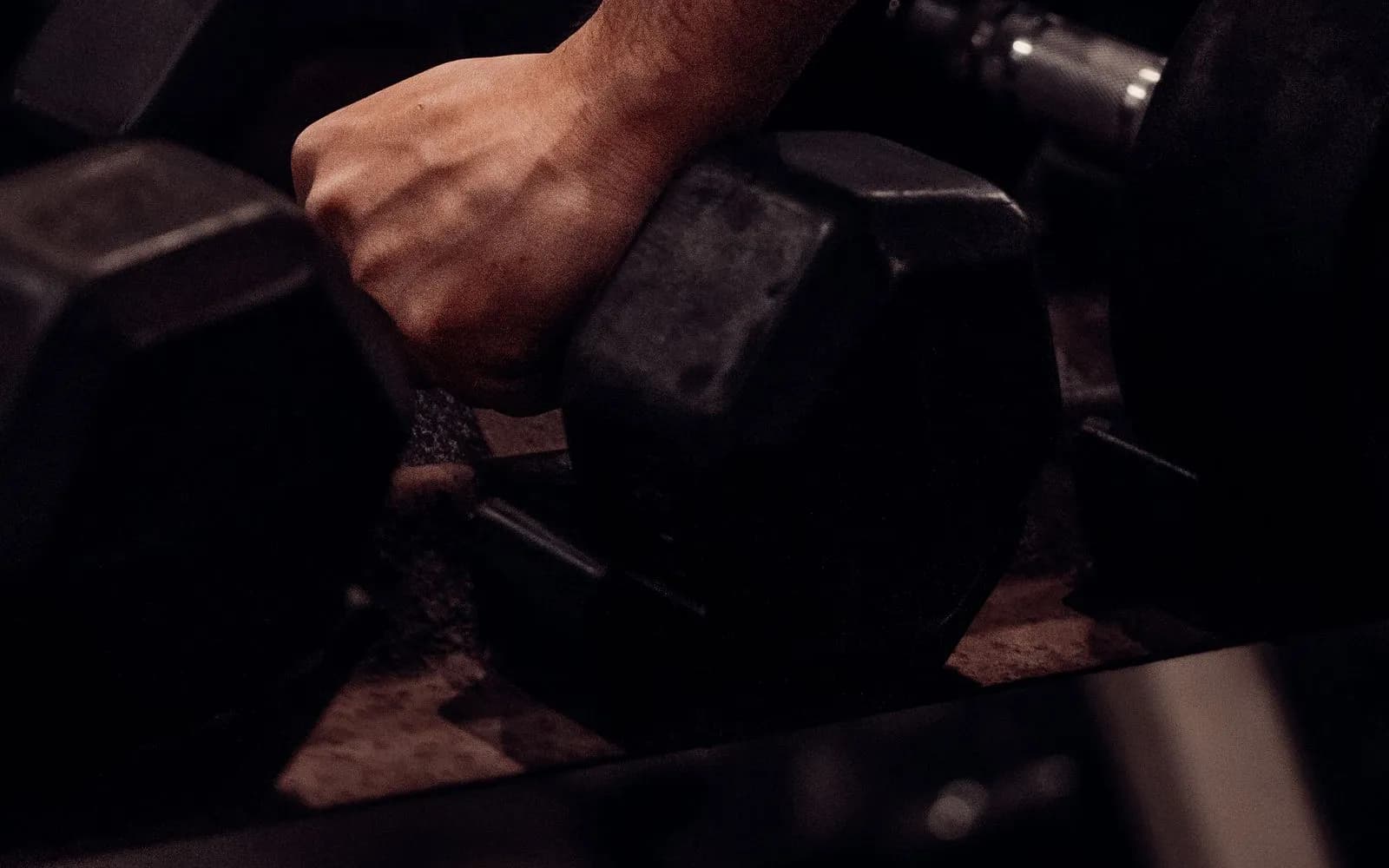 A hand gripping a black hex dumbbell on a rack, with a blurred gym behind