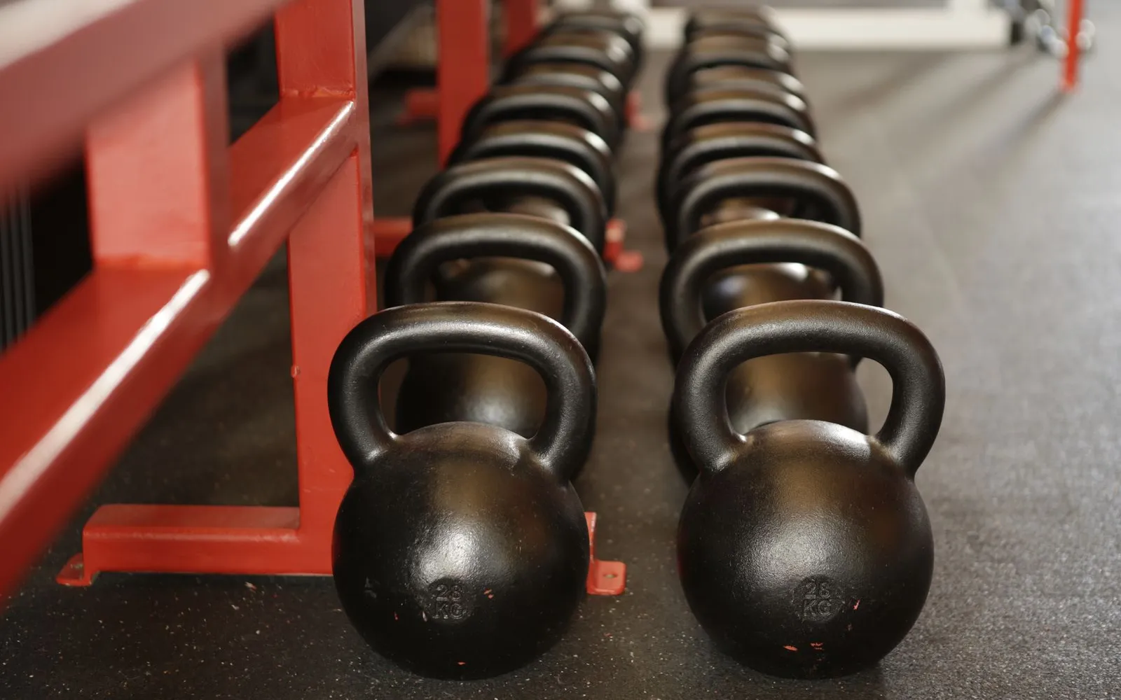 Two rows of black kettlebells lined up on a gym floor beside a red rack