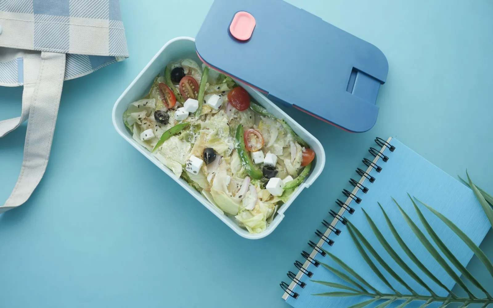 A lunch box of salad with lettuce, cherry tomatoes, olives, green pepper and cubes of cheese, next to a notebook on a blue table
