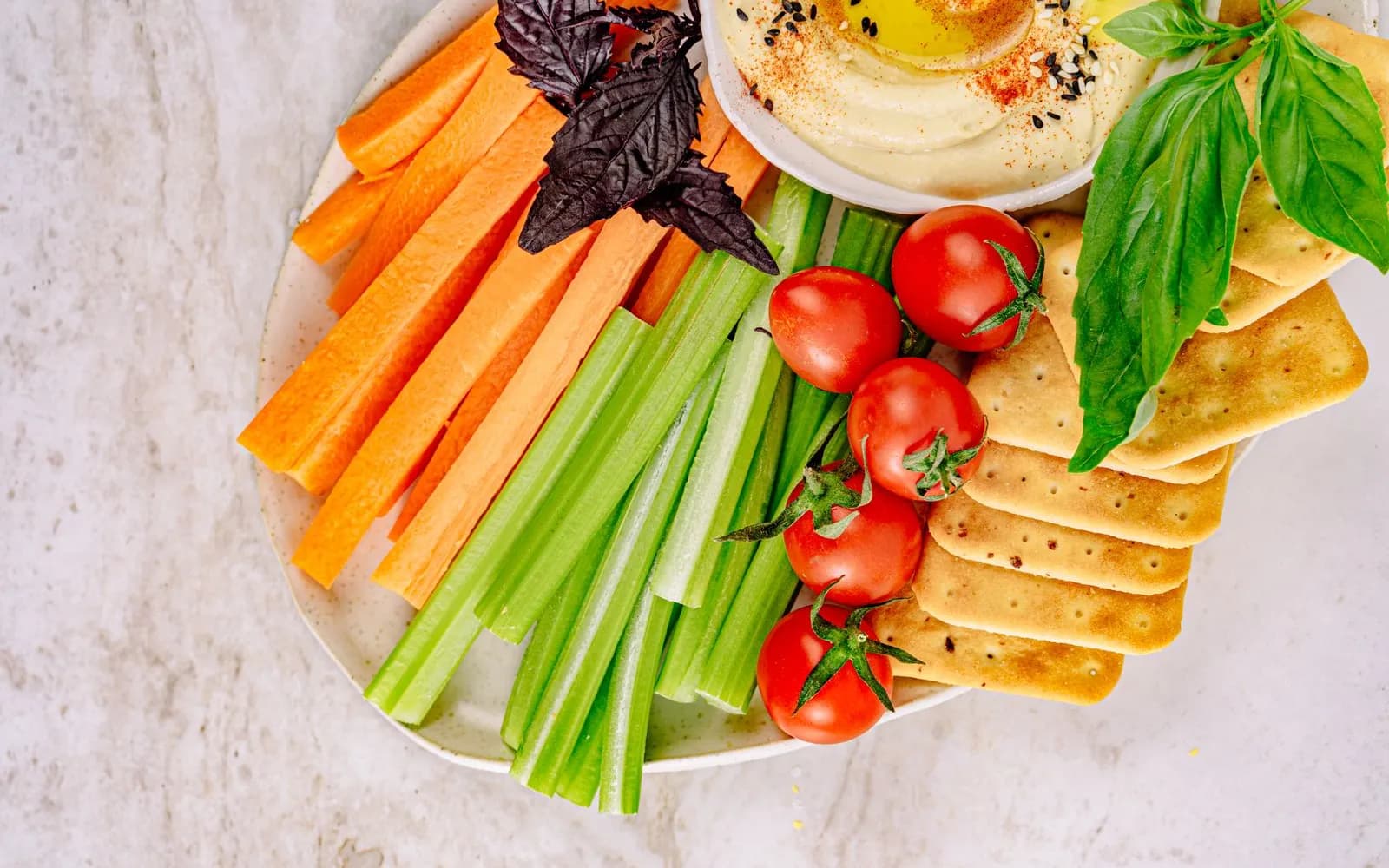 A plate of carrot and celery sticks, cherry tomatoes and crackers with a bowl of houmous