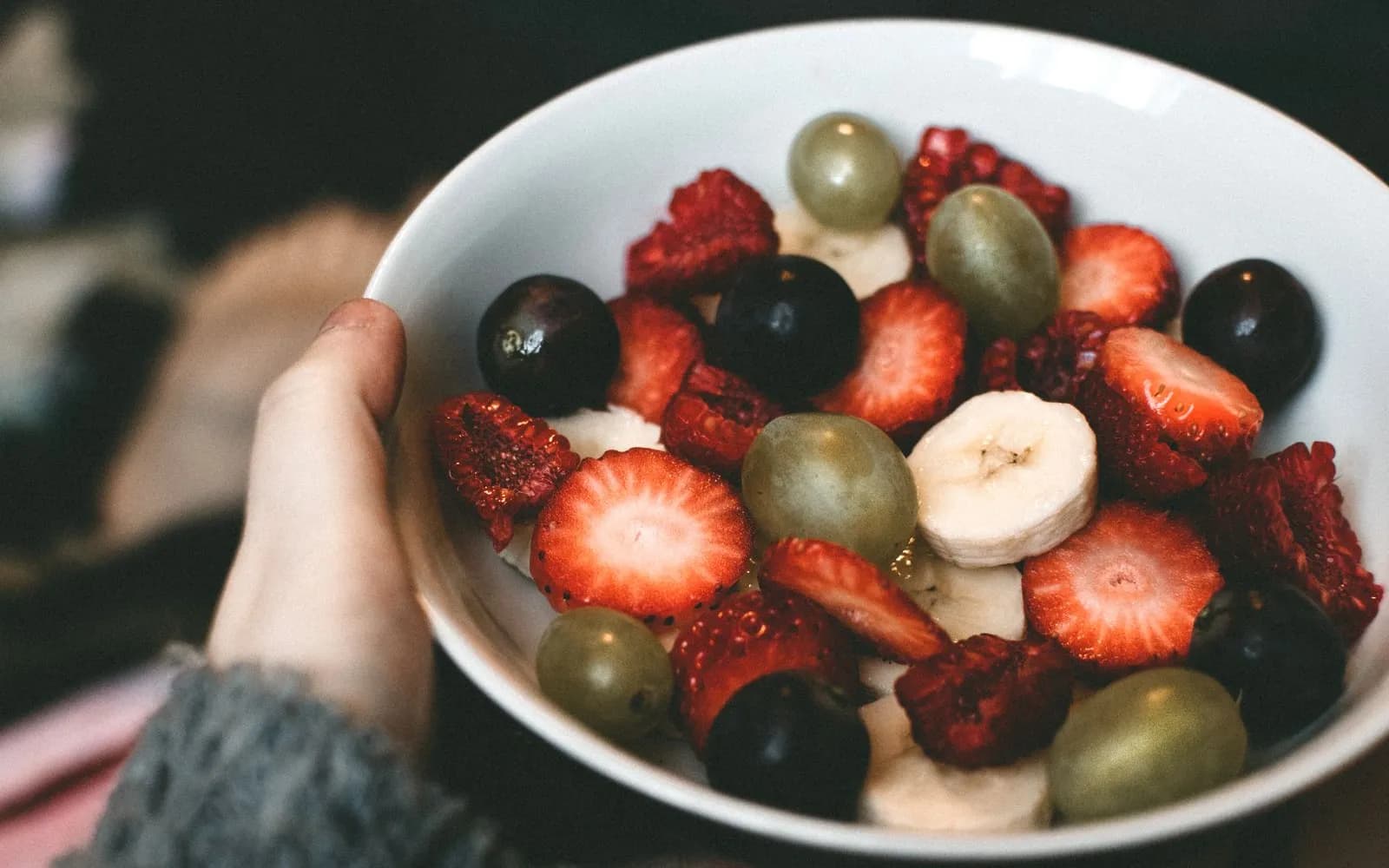 A hand holding a white bowl of sliced strawberries, raspberries, grapes and banana