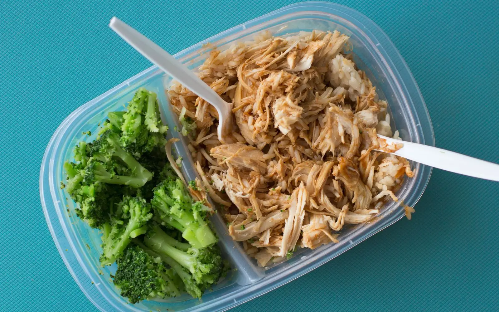 A clear plastic meal prep box with broccoli in one side and shredded chicken on rice in the other, with a plastic fork