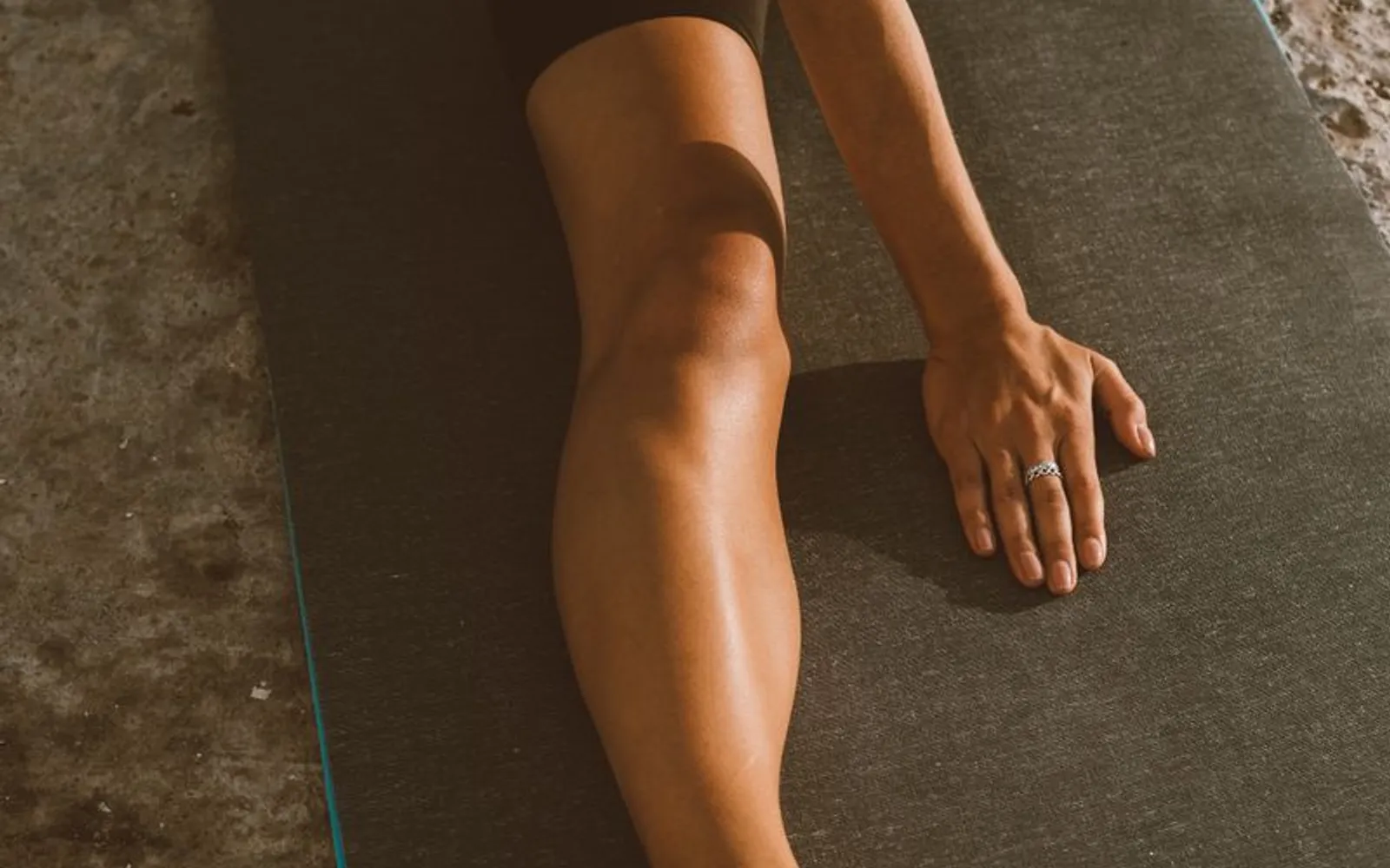 A person's leg stretched out along a dark yoga mat in a low hip stretch, with one hand resting on the mat