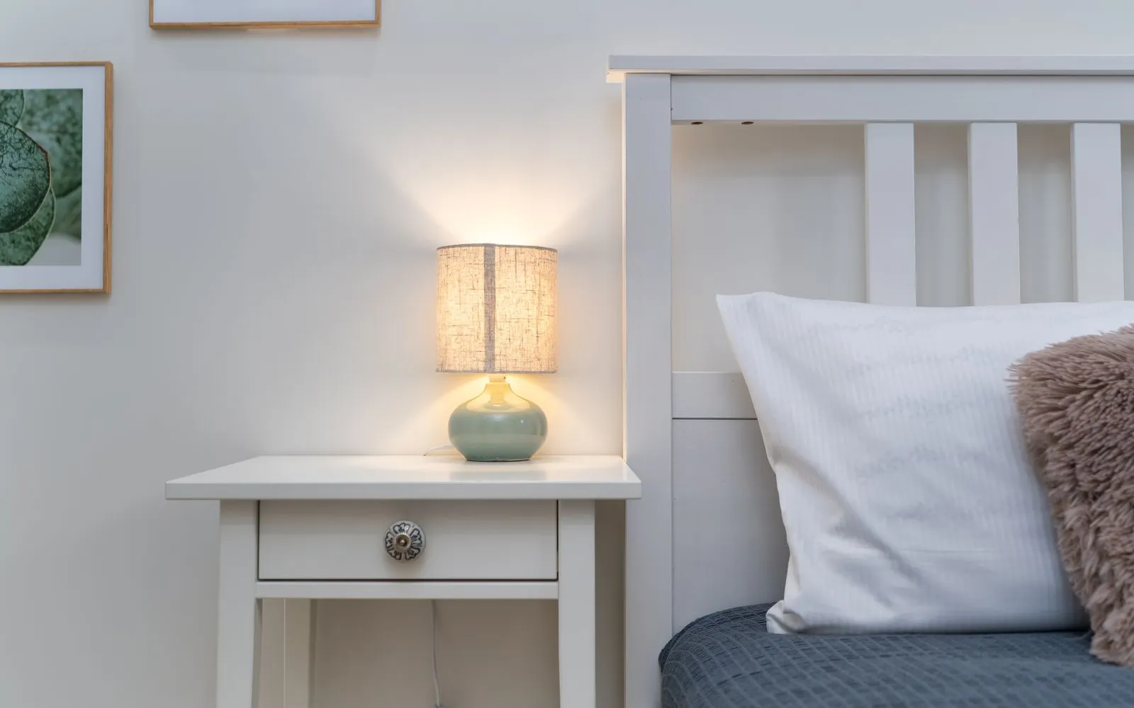A lit lamp on a white bedside table next to a bed with a white pillow and a grey-blue cover