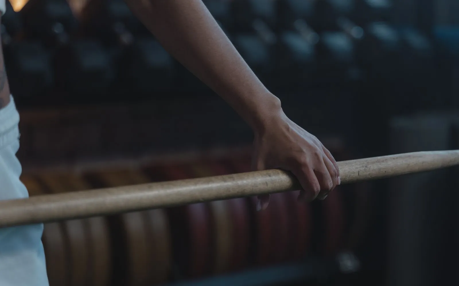 A hand gripping a wooden training bar in a gym, with weight plates blurred behind.