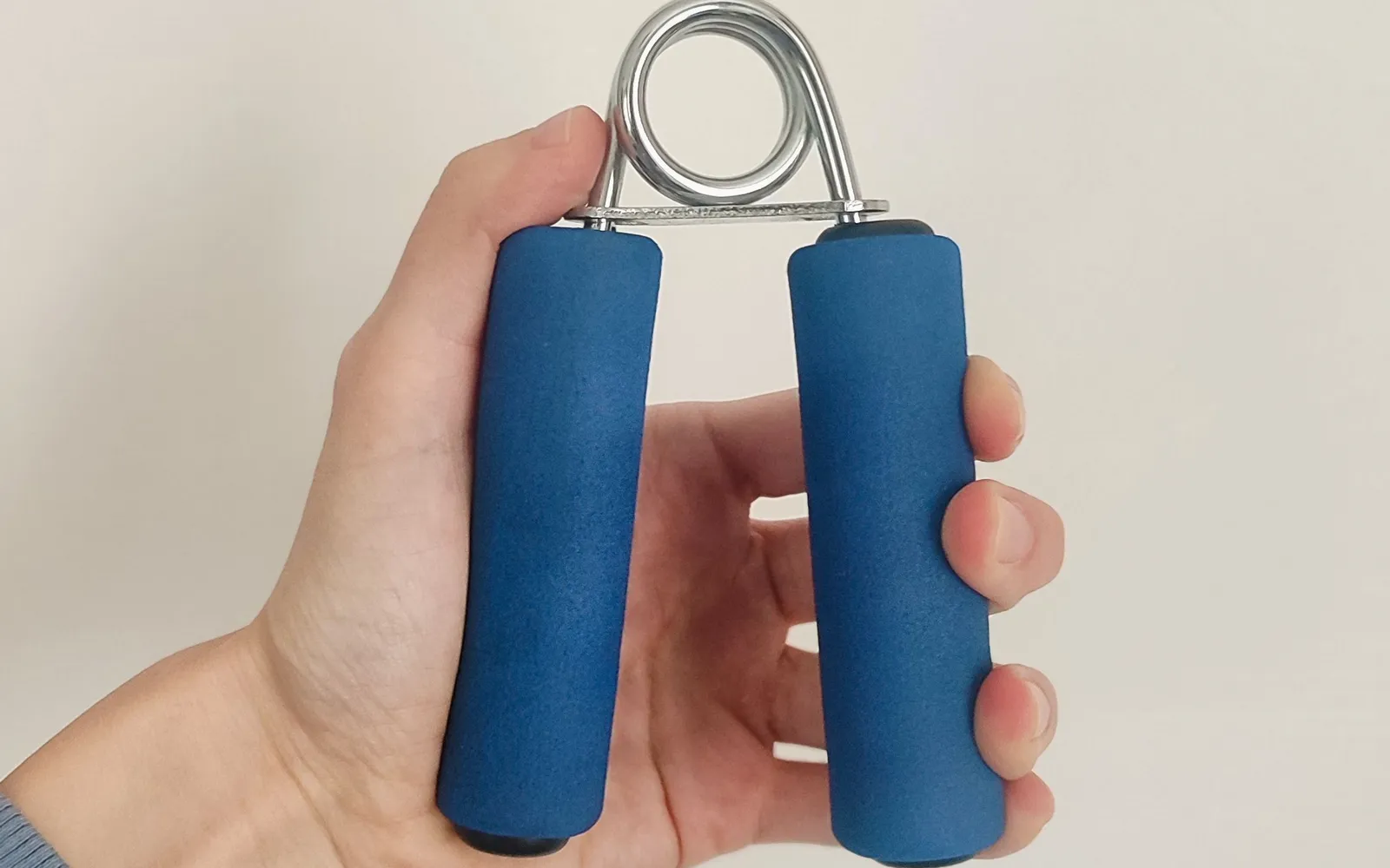 A hand holding a blue foam-handled spring hand gripper against a plain pale wall