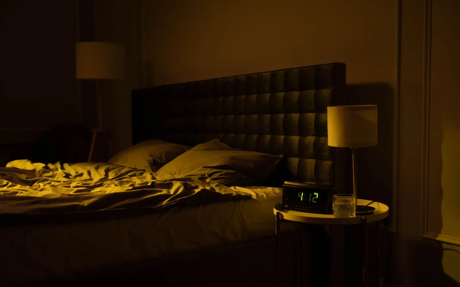 A dim bedroom at night with an empty, unmade bed and a digital clock showing 4:12 on the bedside table.