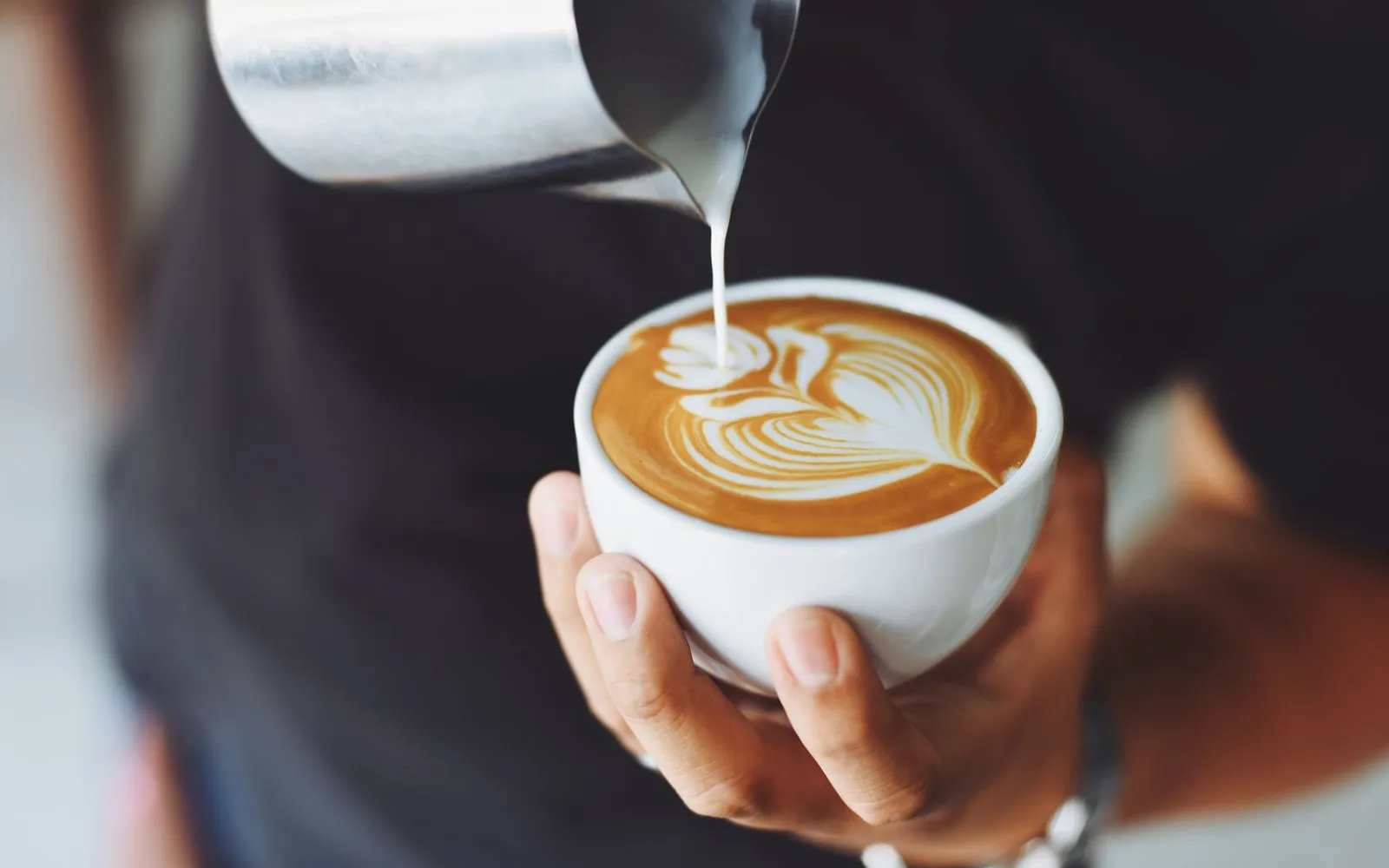 Milk being poured from a steel jug into a cup of coffee held in a hand.