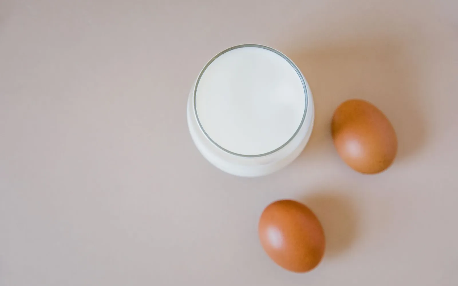 A glass of milk seen from above beside two brown eggs on a beige surface