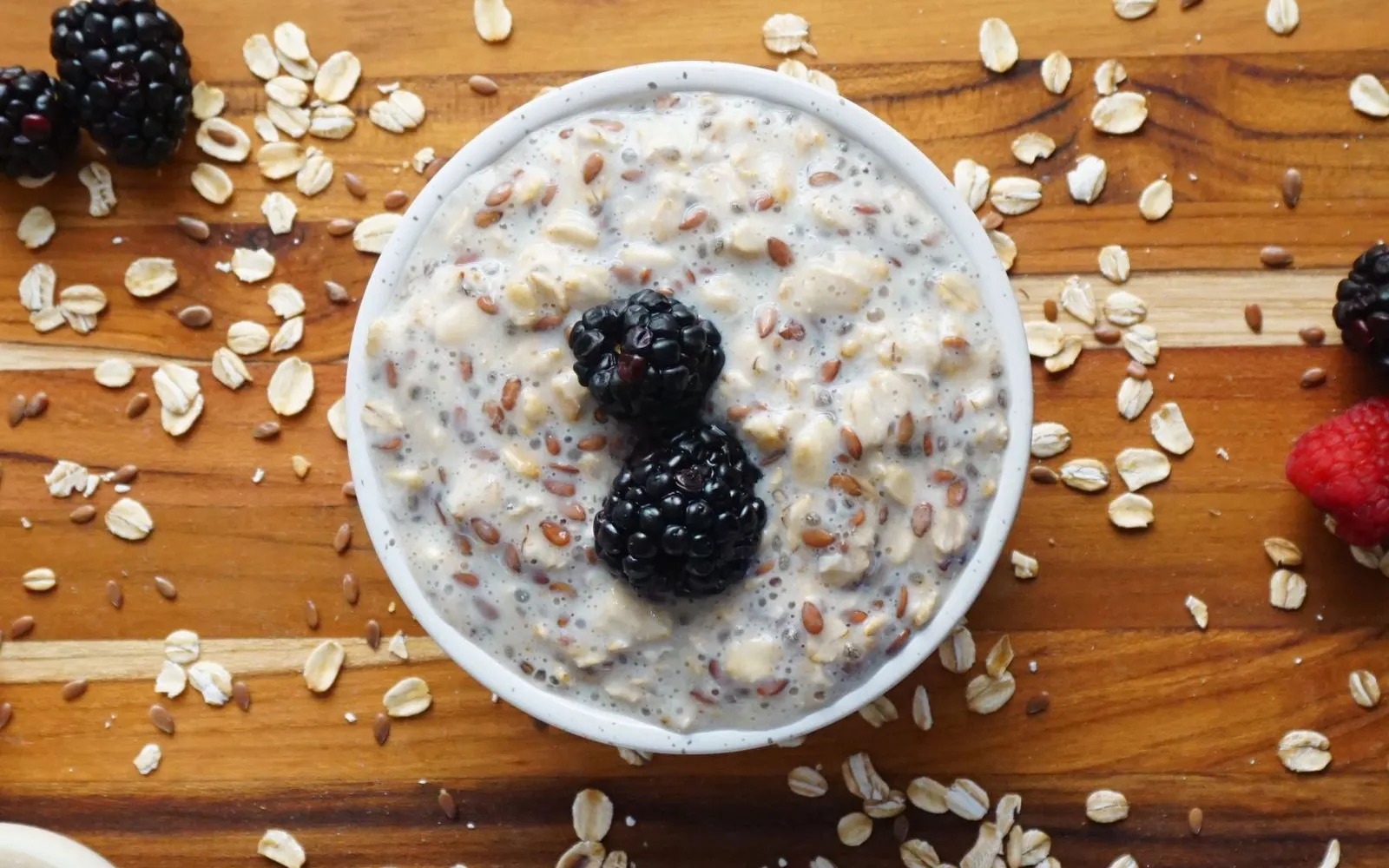 A bowl of overnight oats with flax seeds, topped with two blackberries, on a wooden board scattered with oats and berries