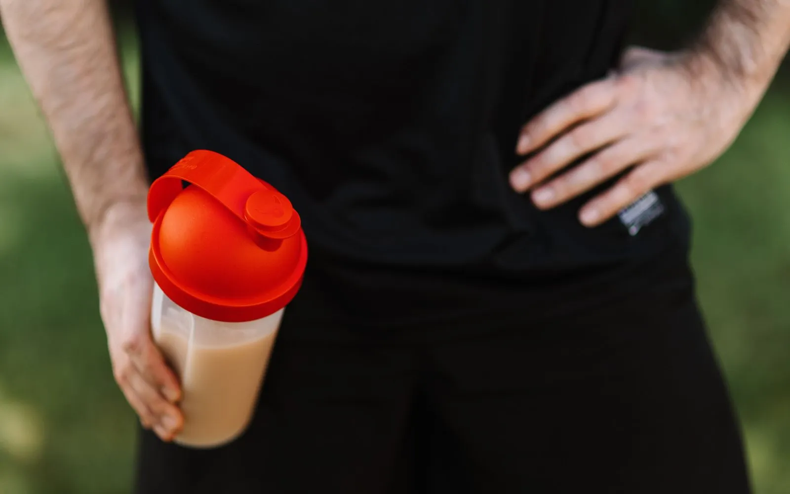 A hand holding a shaker bottle of protein shake with a red lid, beside a person in black sportswear; no face in shot