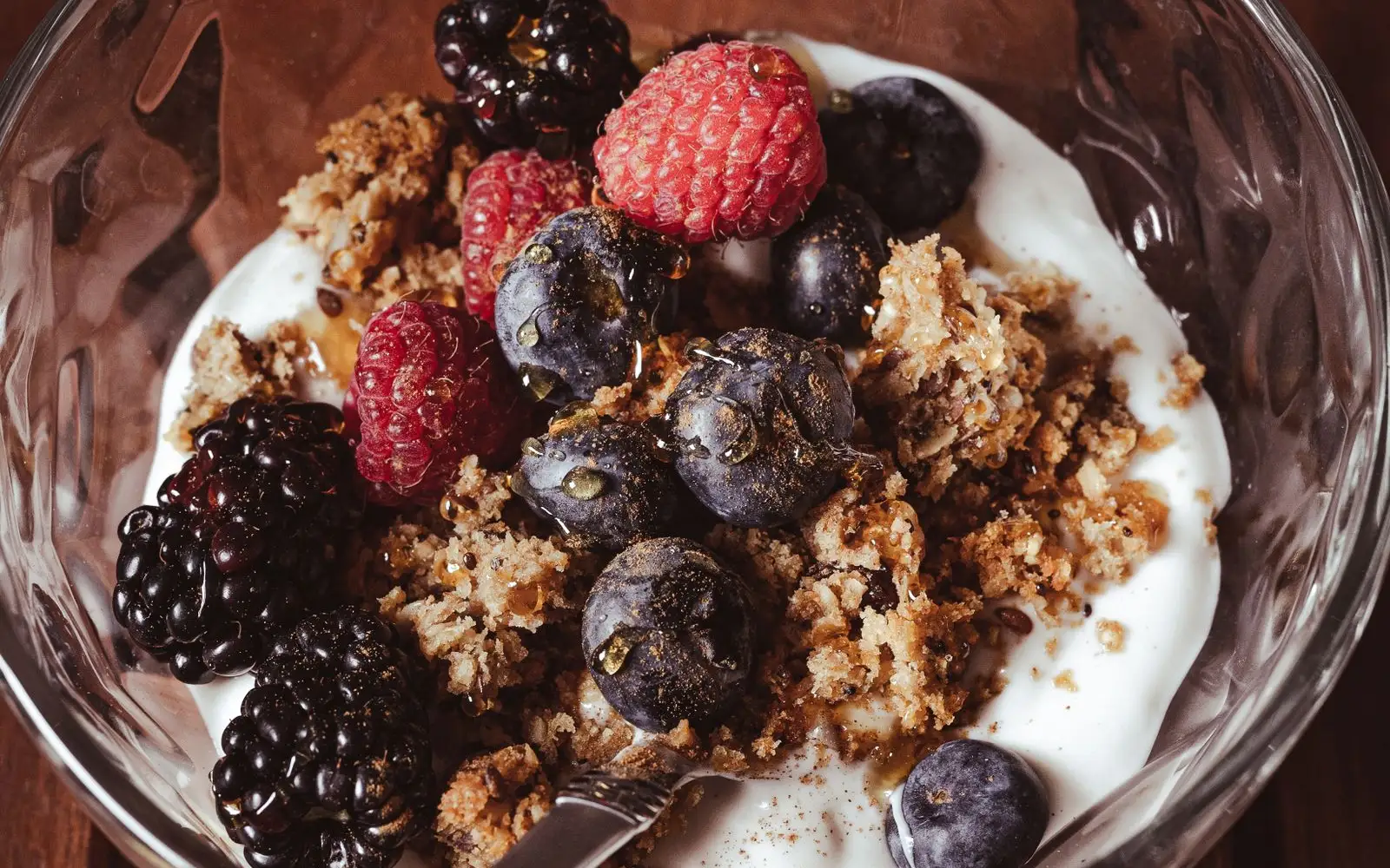 A glass bowl of plain white yoghurt topped with blackberries, raspberries, blueberries and crumbled granola, with a spoon
