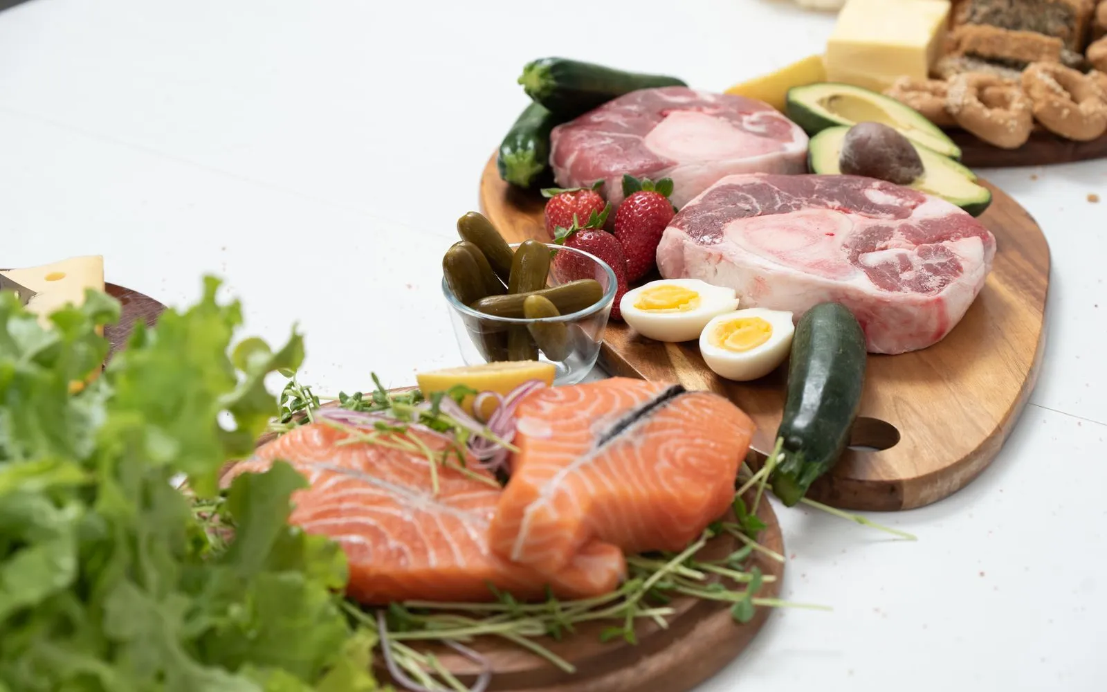 Raw salmon fillets, two raw beef steaks, halved boiled eggs and cheese on wooden boards with strawberries, courgettes and avocado
