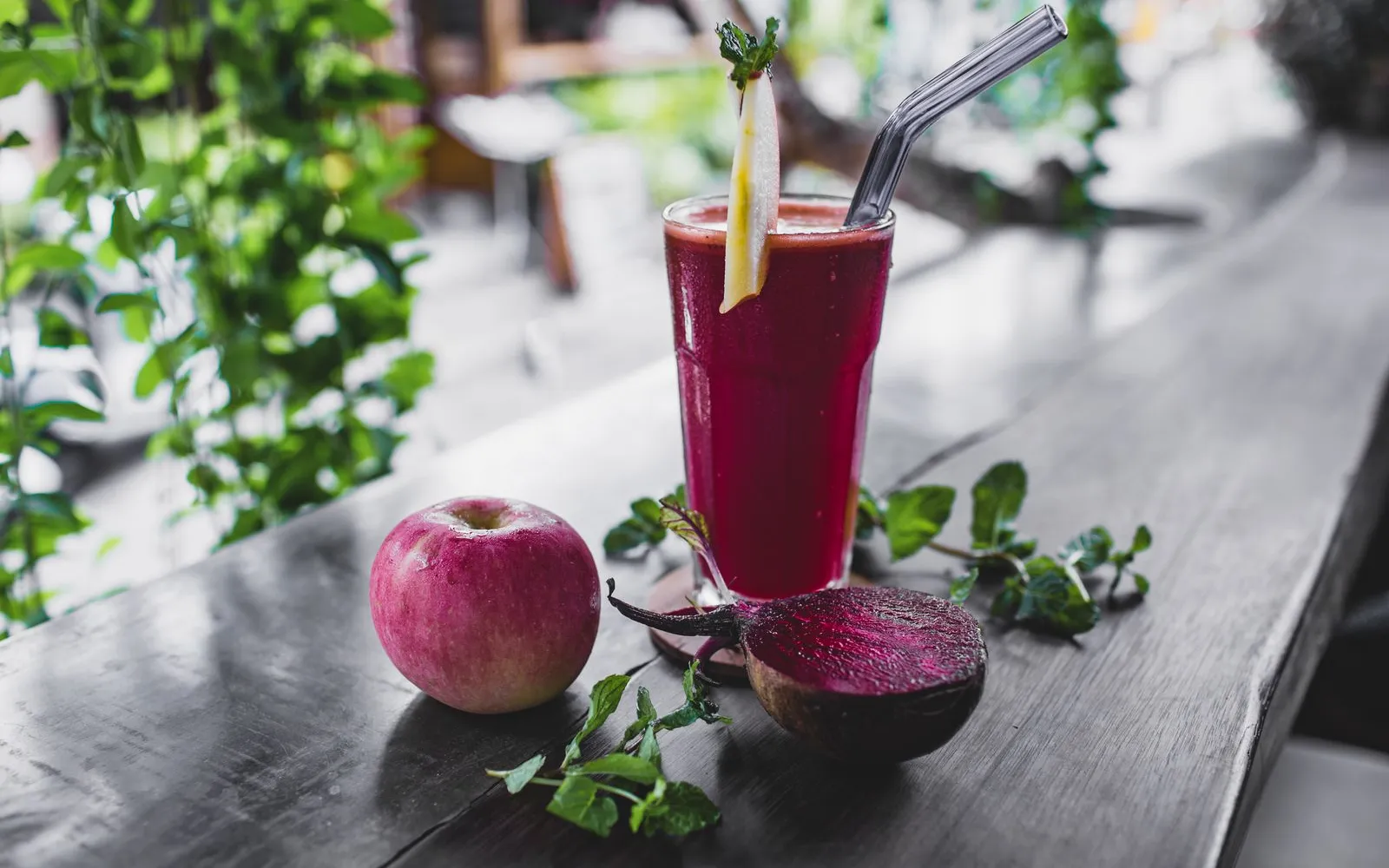 A glass of red beetroot drink with a straw on a wooden table, next to a halved beetroot, an apple and mint leaves