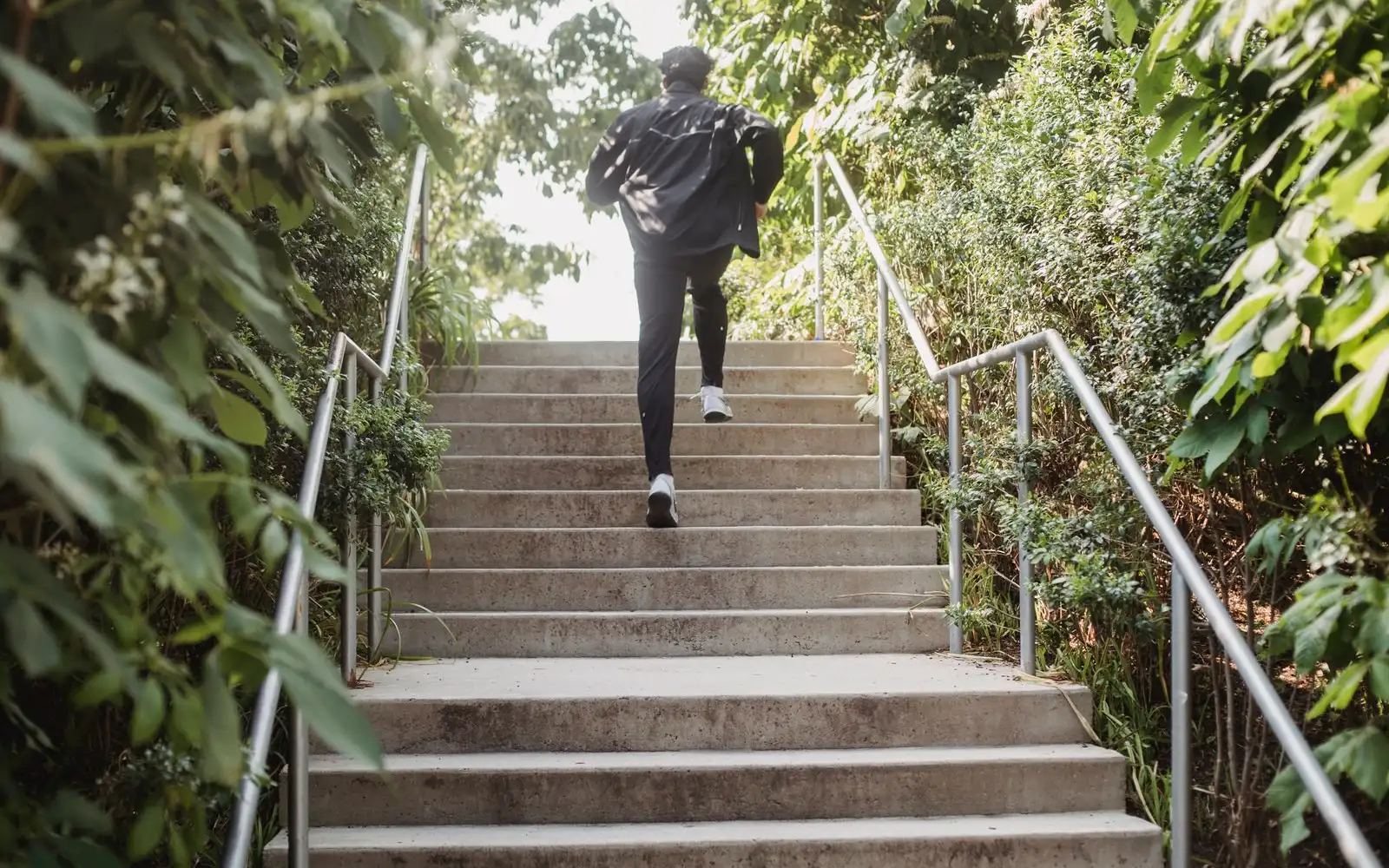 A runner seen from behind running up outdoor concrete steps between green bushes.