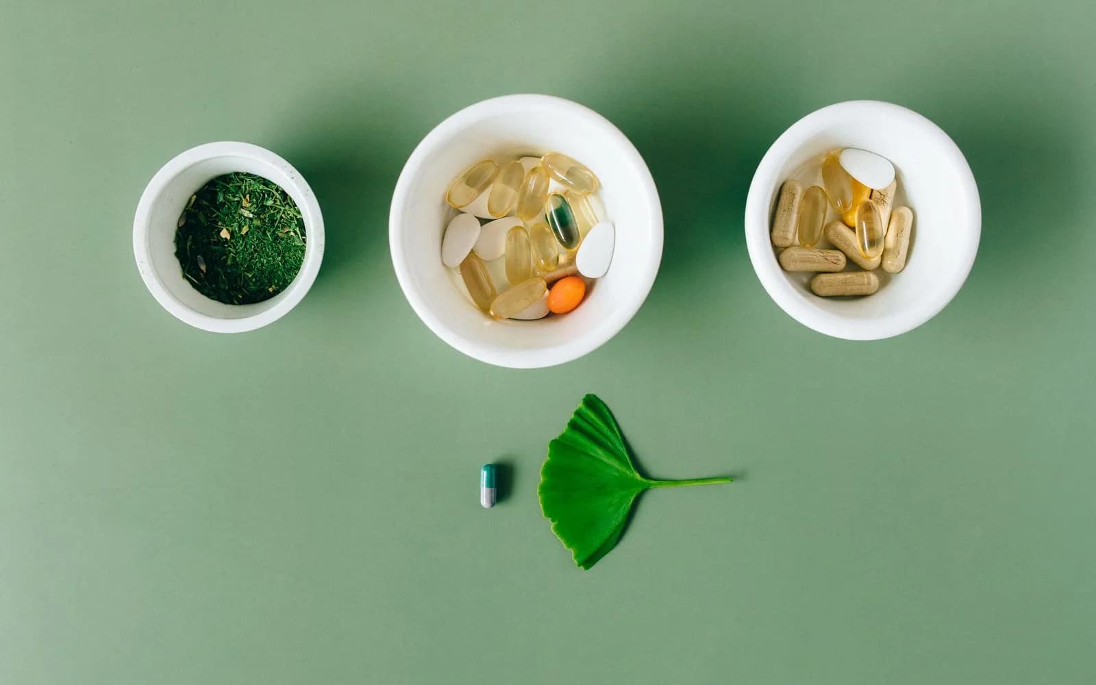 Three white bowls on a green surface holding dried green herbs, soft-gel capsules and herbal capsules, with a single capsule and a leaf below