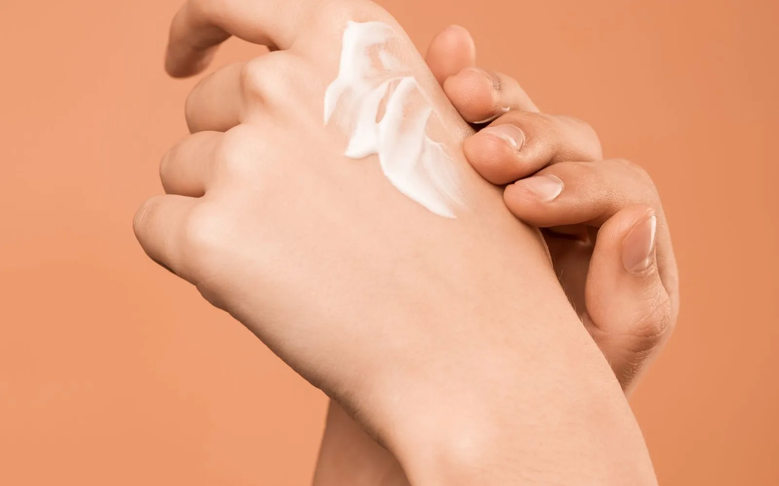 Two hands against a plain peach background, with a swirl of white cream on the back of one hand.