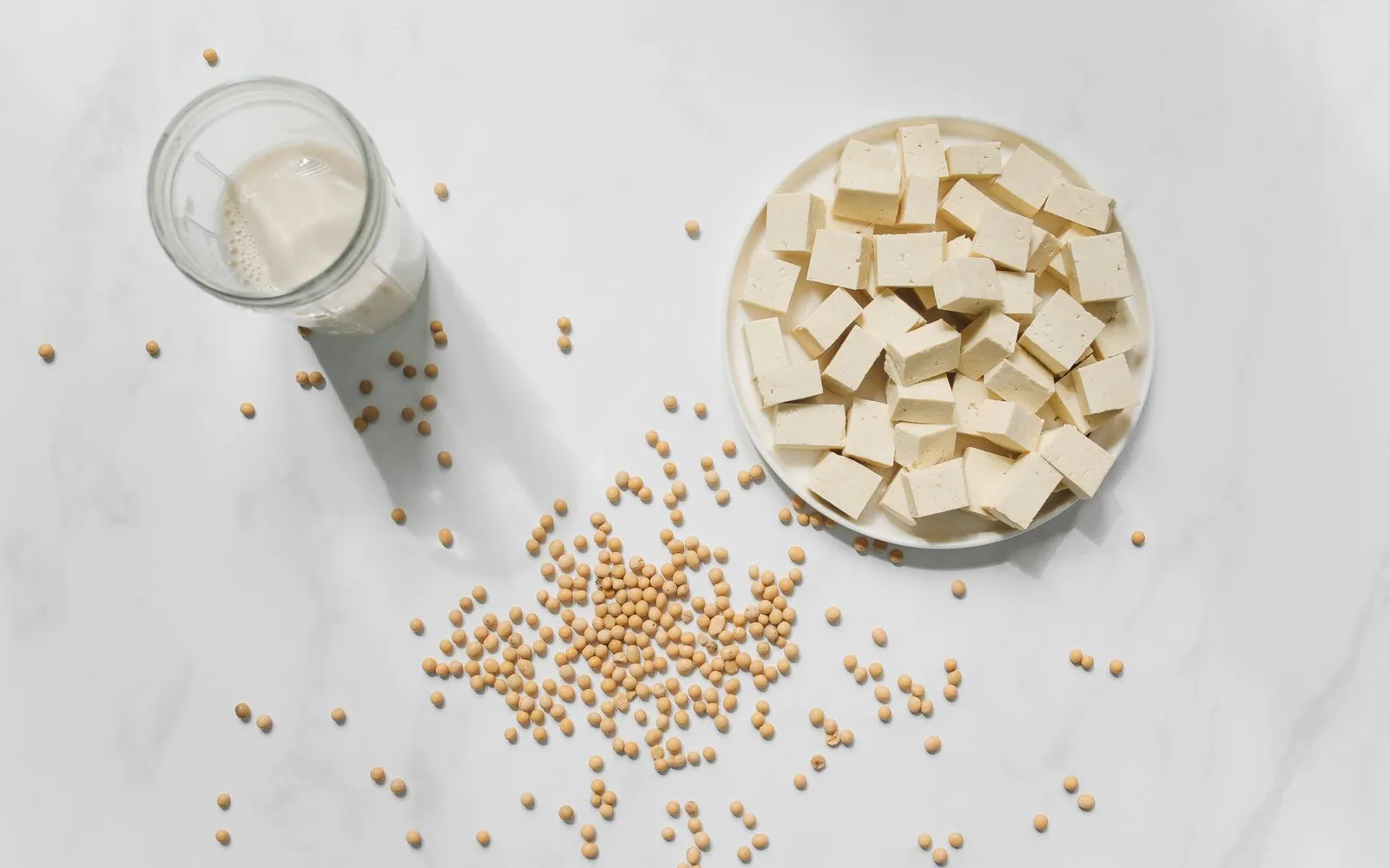Cubes of tofu on a white plate, a glass of soya drink and scattered dried soya beans on a white marble surface, seen from above