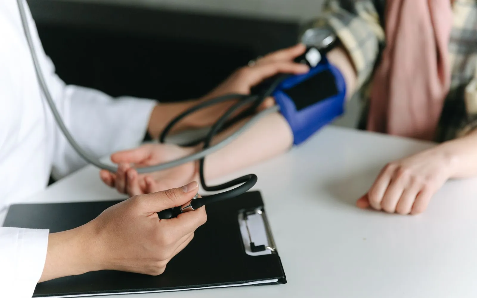 A clinician in a white coat checks a seated person's blood pressure with an arm cuff and hand pump at a white desk.